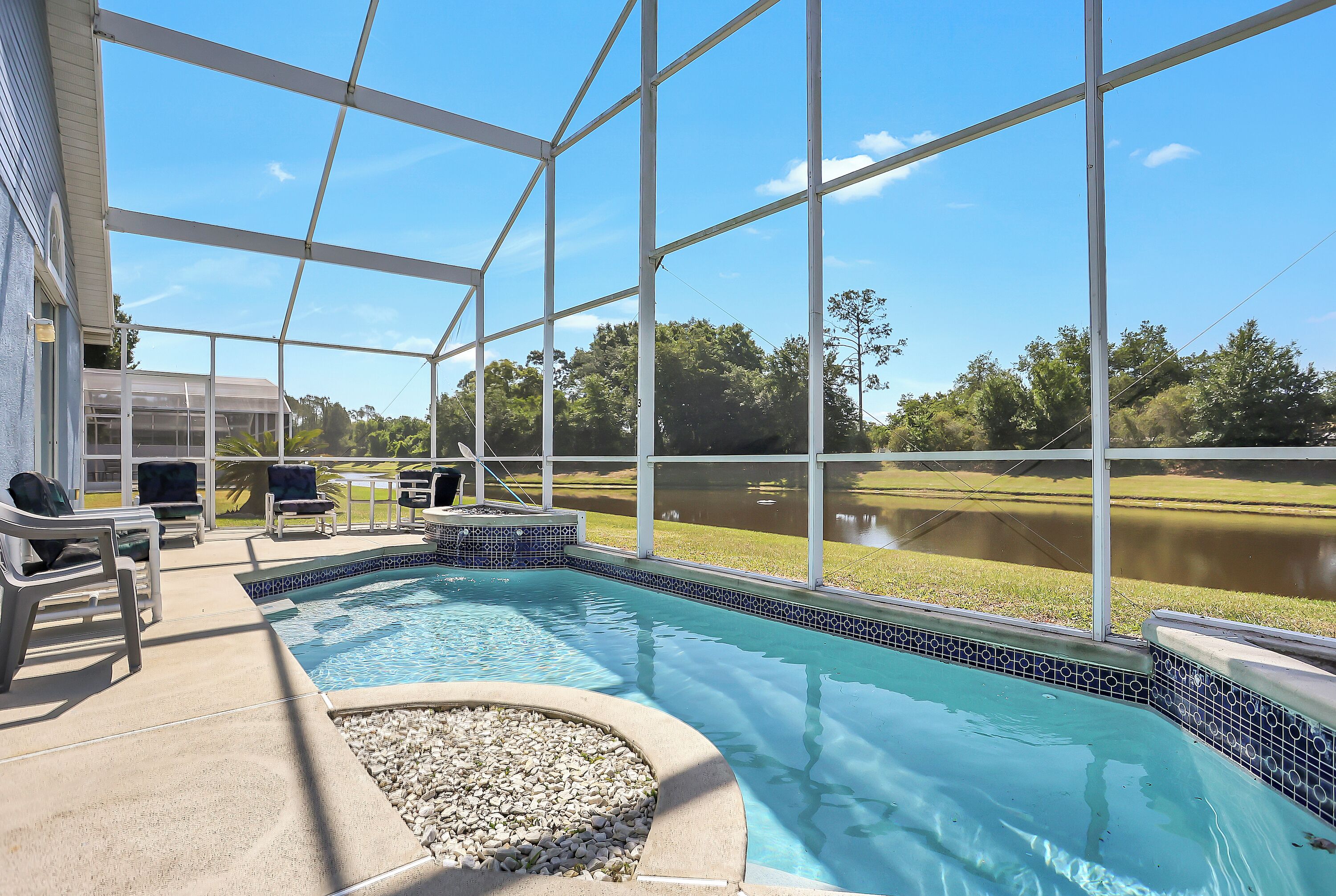 A swimming pool with a view of a lake, surrounded by a covered patio area with chairs and a barbecue grill.