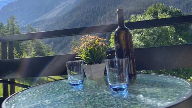 A glass table with a bottle and two glasses, set against a mountain backdrop.