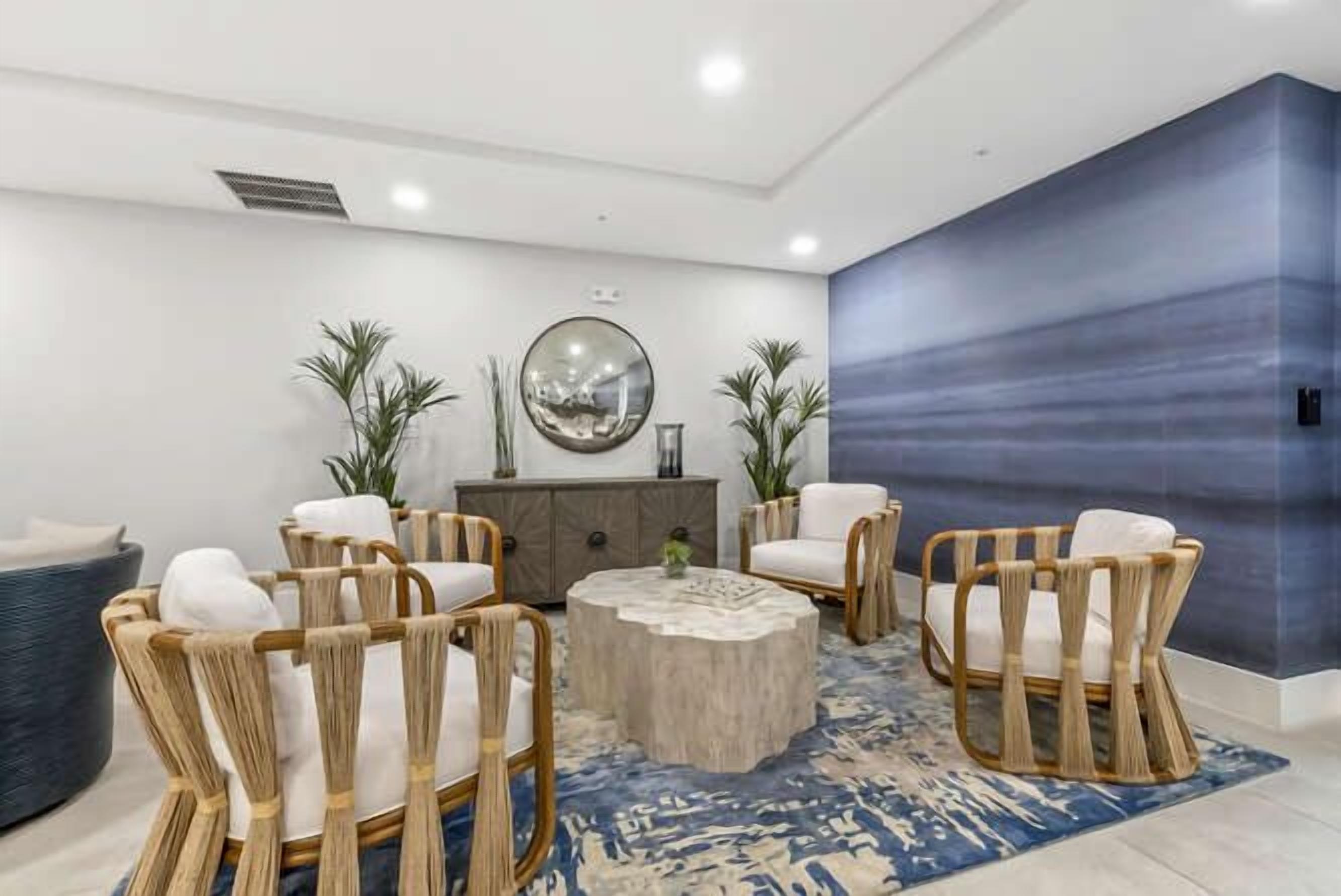 A modern living room with a round coffee table, wicker chairs, a blue patterned rug, and a large mirror on the wall.