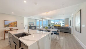 A modern kitchen with a large island, a dining area with a table and chairs, a living room with a sofa, and large windows offering a view of the ocean and buildings. - Gorgeous Third Floor Luxury Residence with Beach Views: Residence 310 (Siesta Key)
