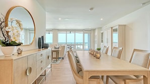 Un comedor amplio con una mesa larga y sillas, un espejo grande y vista al mar a través de amplias ventanas.