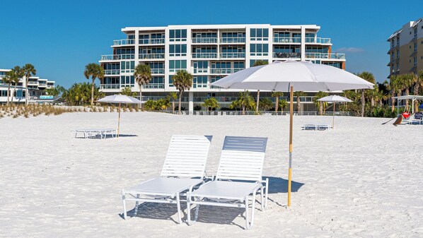 A beach with white lounge chairs and umbrellas, a modern building in the background, and palm trees.