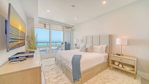 Ein modernes Schlafzimmer mit einem großen Bett, einem Schreibtisch mit Laptop, einem Fernseher und einem Balkon mit Meerblick.