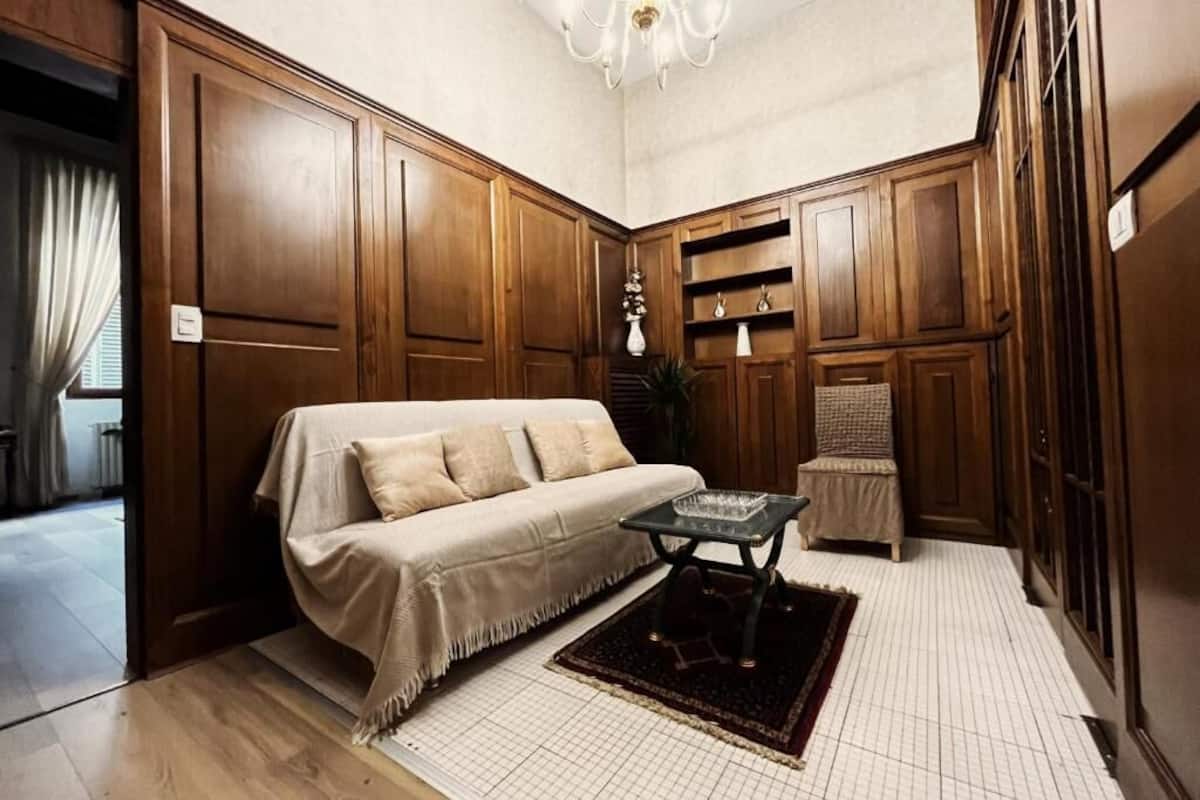 Una habitación con un sofá color beige, una mesita pequeña y una silla. Se observan armarios de madera y un candelabro.
