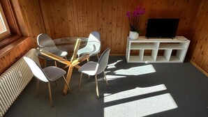 Quarto com parede de madeira, mesa de vidro com cadeiras, suporte branco para TV e um radiador.