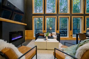 A cozy interior with a fireplace, a large window with a forest view, and comfortable seating.