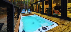 A hot tub on a wooden deck with lounge chairs, a dining area, and a kitchen visible through large windows.