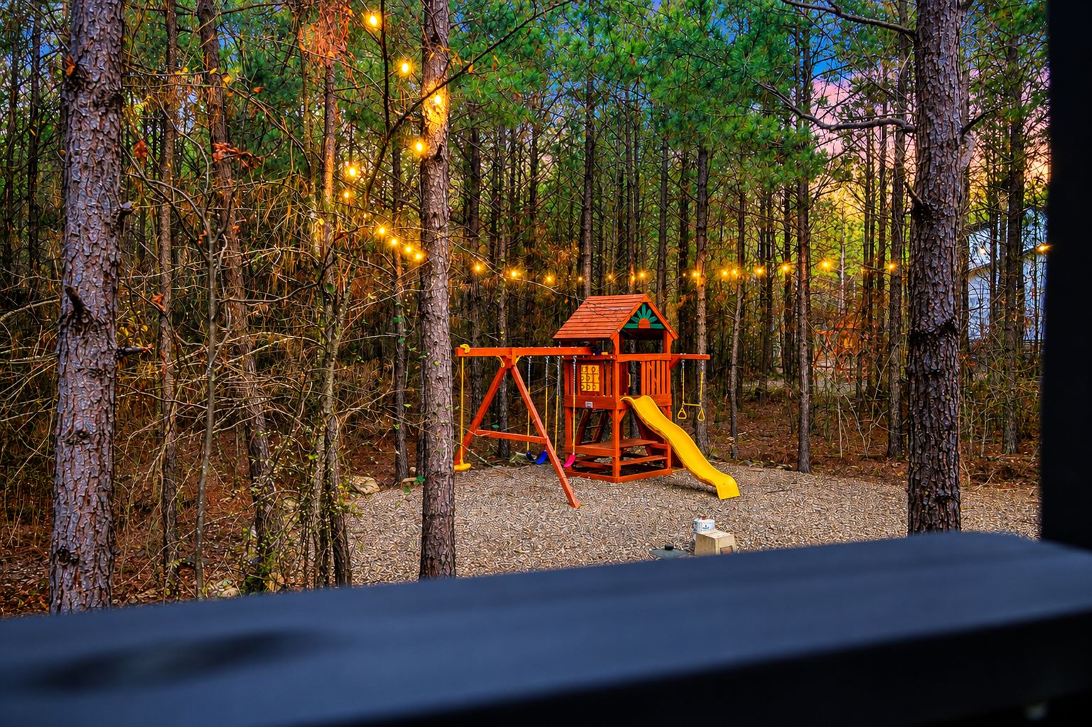 A playground with a slide and swings, surrounded by trees and string lights.