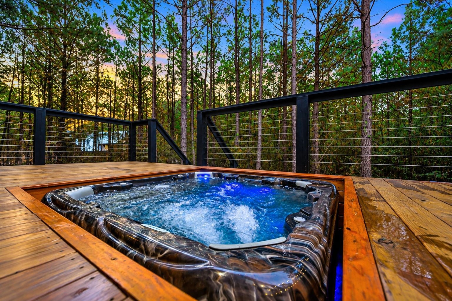 Ein Whirlpool auf einer Holzterrasse, umgeben von einem Metallgeländer, mit einem Wald im Hintergrund.