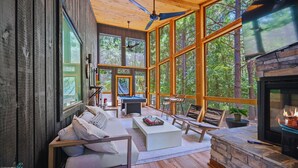 A spacious living room with a stone fireplace, large windows, and a ceiling fan. - Swim & Recharge | Heated Pool Spa | Pool Table (Broken Bow)