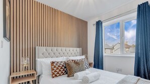 Un dormitorio con cabecera capitonada, pared de listones de madera, un ventanal con cortinas azules y una cama bien hecha con almohadones estampados.