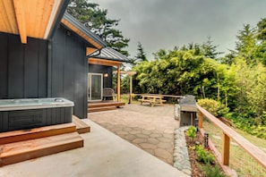 A patio area with a hot tub, picnic table, and barbecue grill, surrounded by a wooden deck and stone paving.