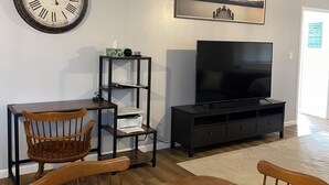 A living room with a wooden dining table, chairs, a television, a bookshelf, and a wall clock.