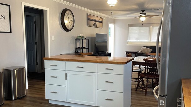 A modern kitchen with white cabinetry, a wooden countertop, and a stainless steel refrigerator. There is a dining area with a table and chairs, and a ceiling fan above.