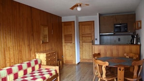 Een gezellige kamer met houten vloer, een bank met geruit plaid, een houten eettafel met stoelen, een keukenhoek met een magnetron, en houten deuren.