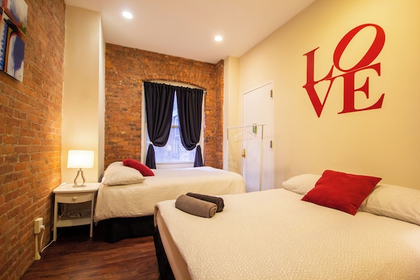 Une chambre d’hôtel avec deux lits, un mur en briques, un grand panneau portant le mot « AMOUR » et une fenêtre avec des rideaux.