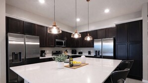 Une cuisine moderne avec des meubles de couleur sombre, des appareils en acier inoxydable, un grand îlot central et des luminaires suspendus.
