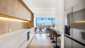 A modern kitchen with a view of the sea, a dining area with bar stools, and a large refrigerator.