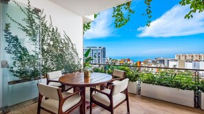 A balcony with a round wooden table and chairs, a glass wall with greenery, and a city view.