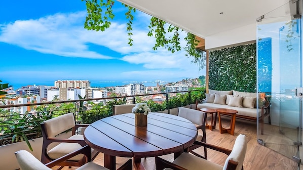 A balcony with a round table and chairs, a sofa, and a view of the city and sea.