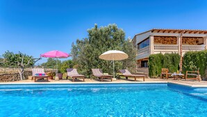 Une piscine avec des chaises longues et des parasols, un bâtiment en pierre doté d’un balcon, et une végétation luxuriante tout autour.