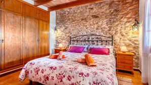 Une chambre avec un mur en pierre, une armoire en bois et un lit agrémenté de draps à motifs floraux et d’oreillers roses.