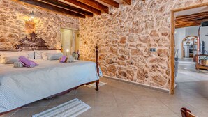 A bedroom with a stone wall, wooden beams, a bed with a patterned bedspread, and a nightstand with books. - Na Corme Nova by dracmallorca (Calonge)