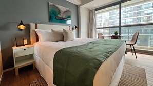 A modern hotel room with a large bed, a desk, and a chair. There is a window with a view of buildings outside.