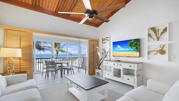 A spacious living room with a ceiling fan, a TV, and a view of the beach through large windows. - New to the rental program! Book your week today! (Captiva)