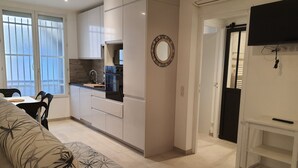 A modern kitchen with white cabinets, a built-in oven, and a microwave. There is a dining area with a table and chairs, and a sofa with a patterned cover. A television is mounted on the wall, and there is a door leading to another room.