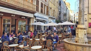 An outdoor café with wicker tables and chairs, a stone fountain, and a building with a sign reading "CAFÉ CLEMENCEAU".