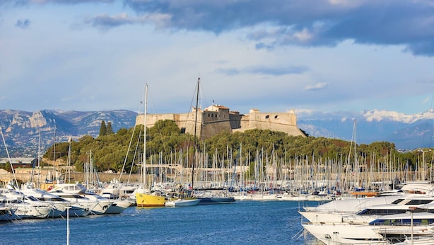 A marina with numerous boats docked, a castle on a hill in the background, and mountains in the distance.