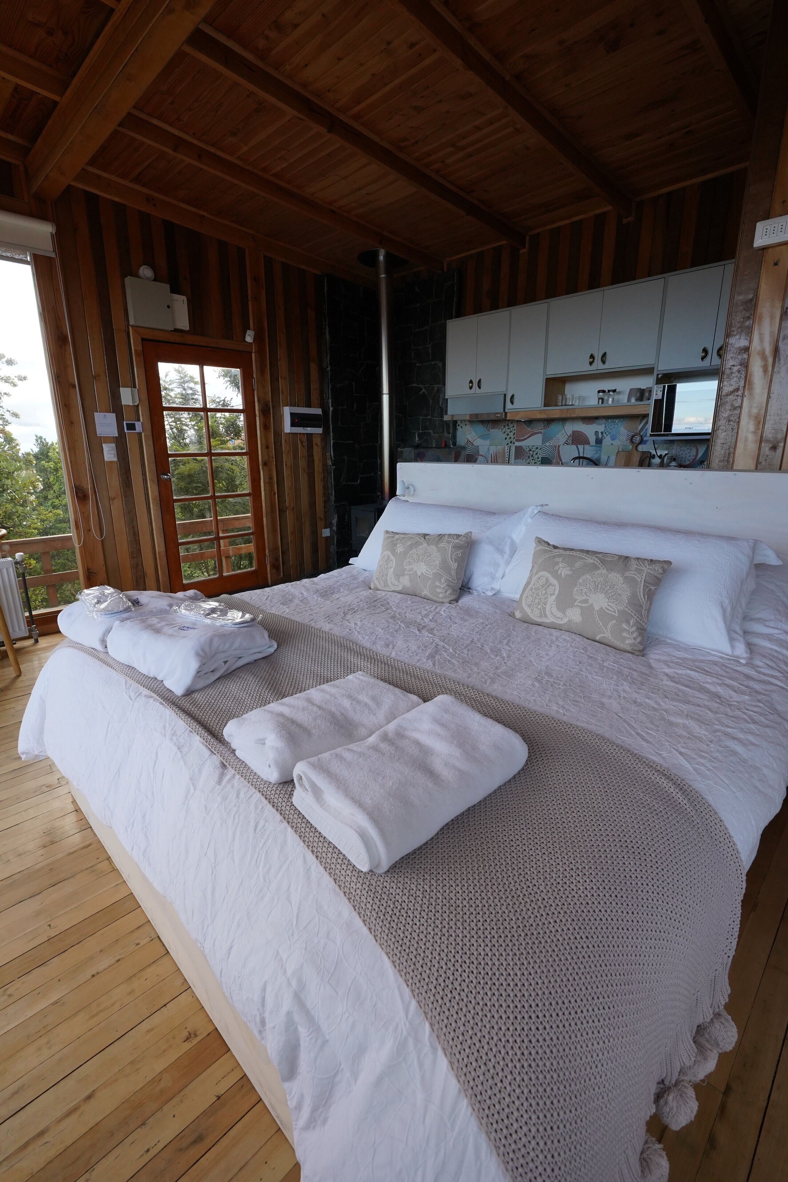 Un dormitorio de cabaña de madera con una cama grande, ropa de cama blanca y vista a la cocina a través de una puerta abierta.