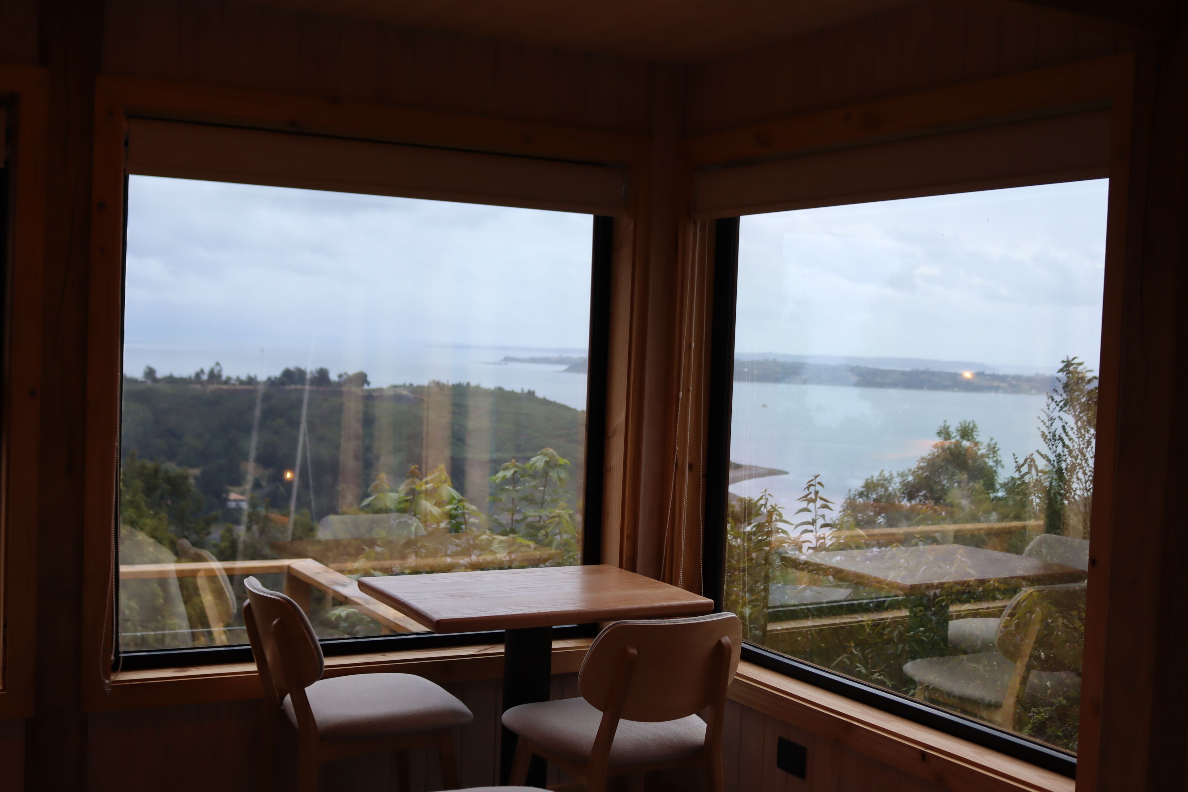A room with a wooden table and two chairs, large windows with a view of a body of water and hills.