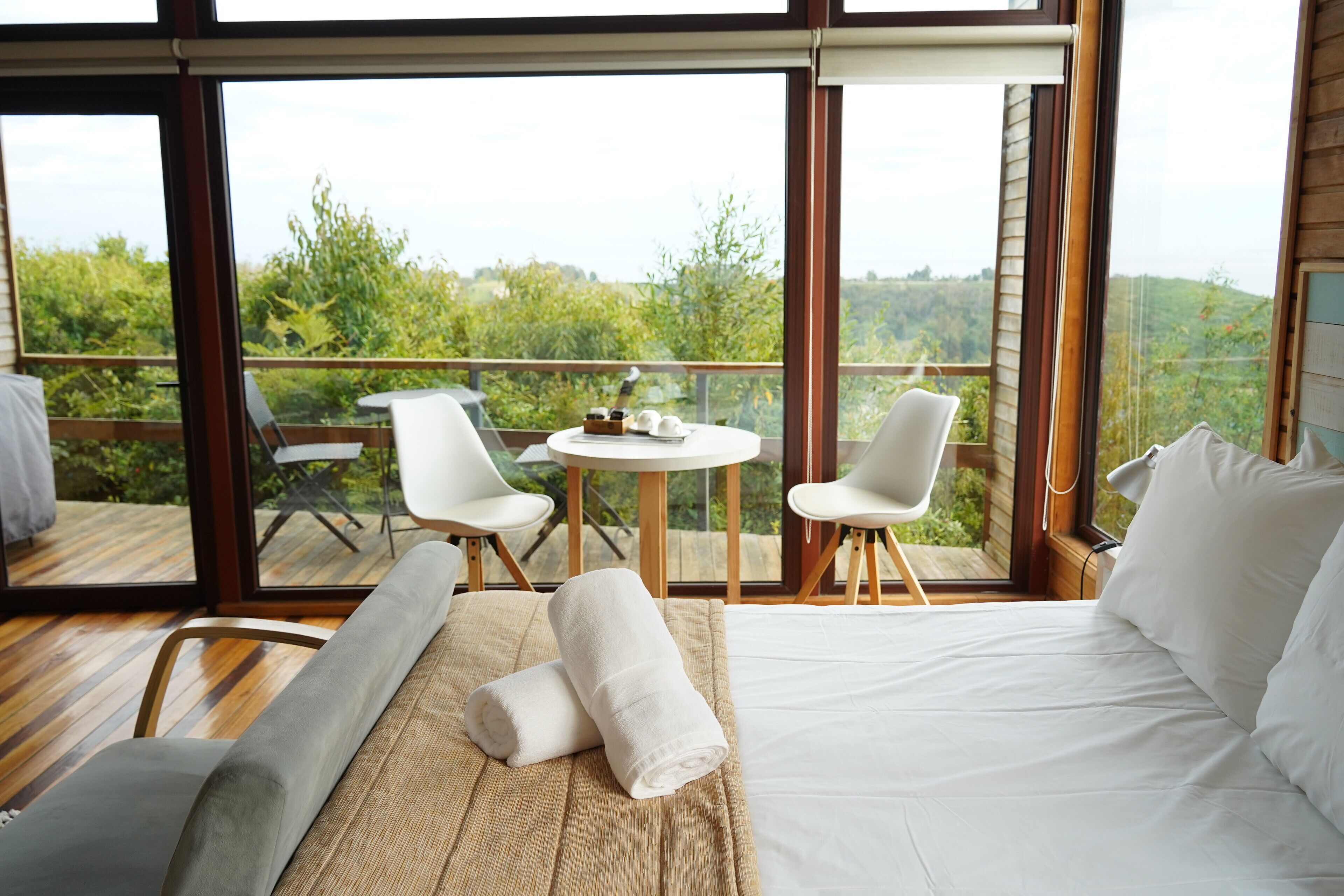 A bedroom with a bed, two chairs, a small table, and a view of greenery through large windows.