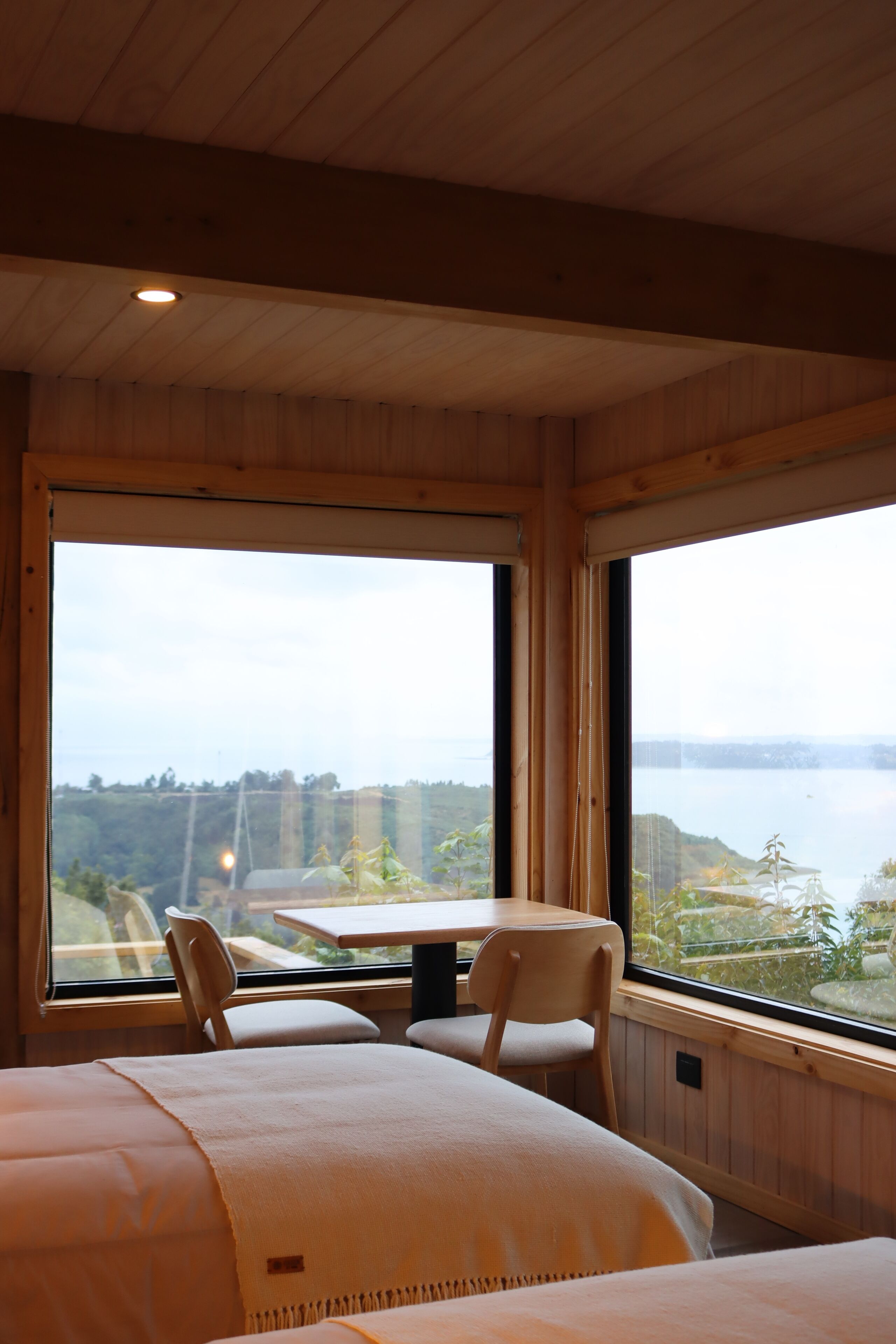 A room with a bed, a table, and two chairs. There are large windows with a view of a body of water and greenery.