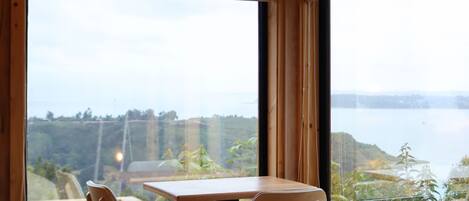 Une pièce comprenant un lit, une table et deux chaises. De grandes fenêtres offrent une vue sur une étendue d’eau et sur la verdure.