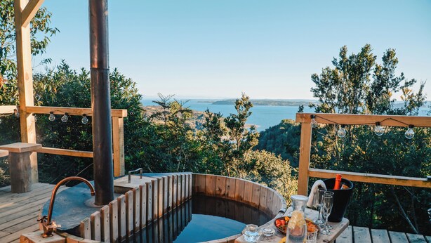 Ein hölzernes Whirlpool auf einer Terrasse mit Tisch und Getränken, mit Blick auf eine reizvolle Landschaft.