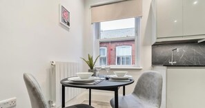 A compact kitchen with a dining table set for two, a window with a view of a red brick building, and a radiator on the wall.
