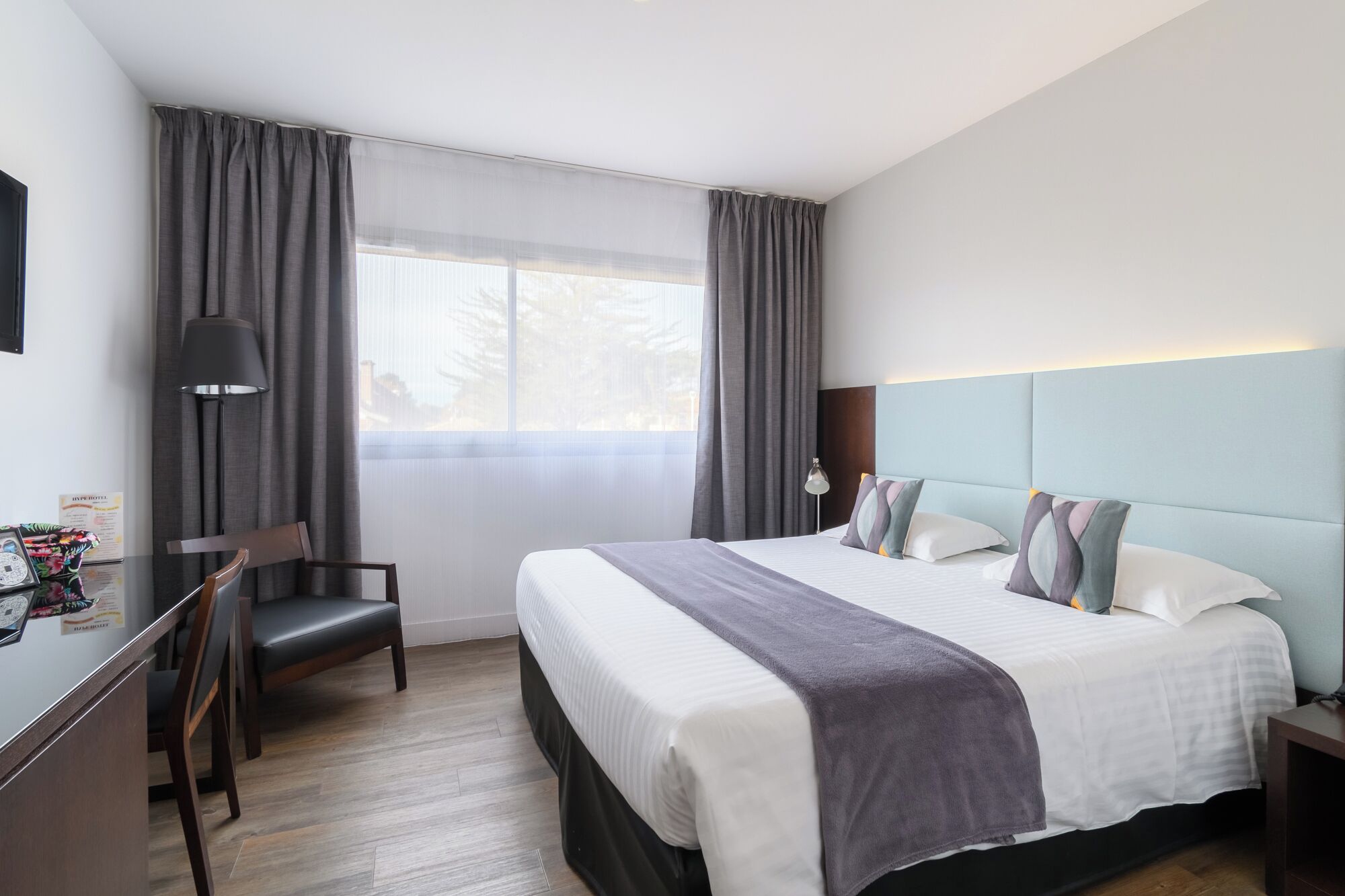 A modern hotel room with a large bed, a desk with a chair, a lamp, and a view of trees through the window.