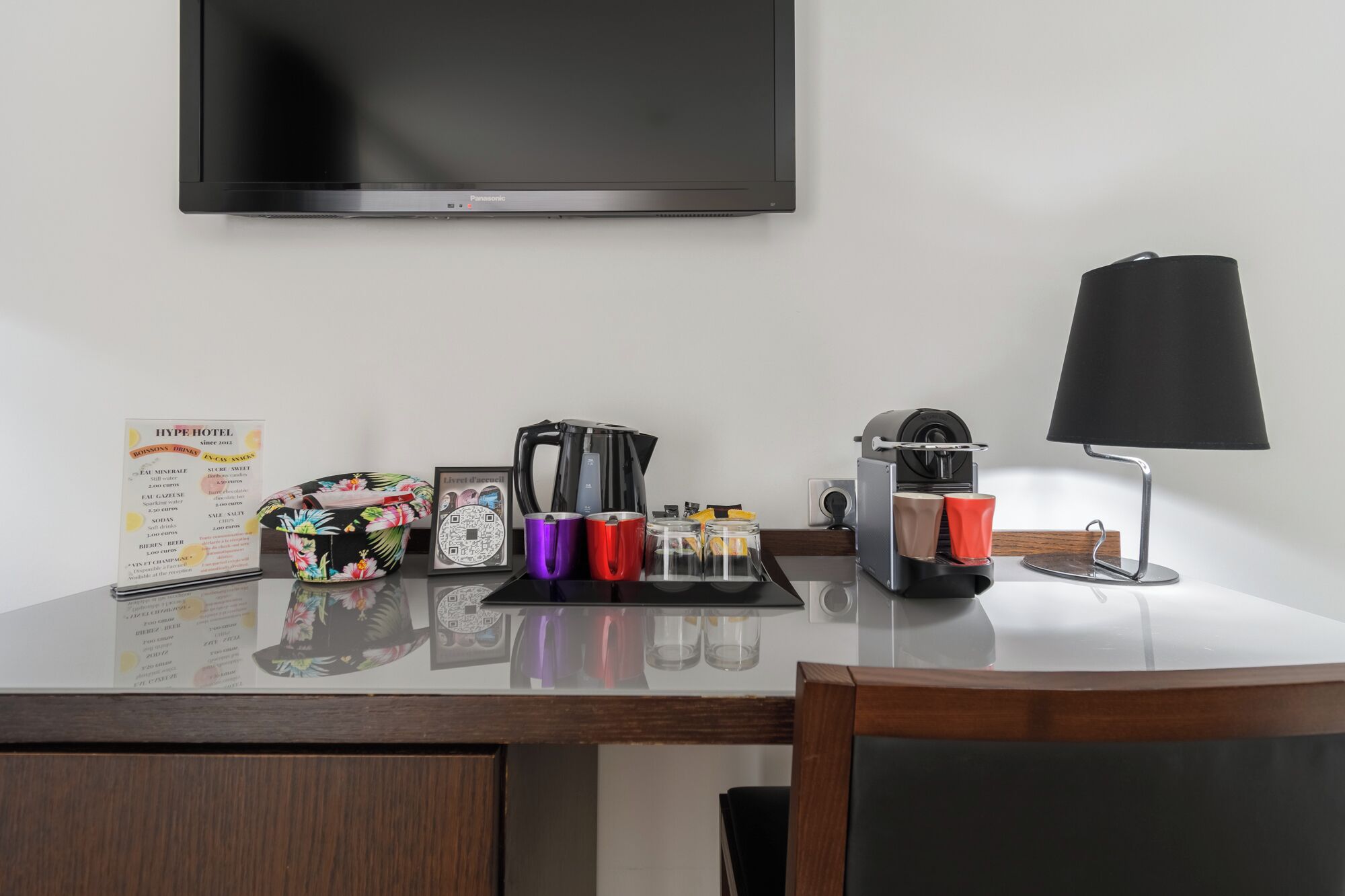 A hotel room desk with a flat-screen TV, a coffee maker, a lamp, a menu, and a floral-patterned bag.