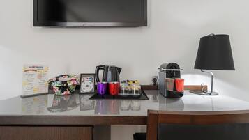 A hotel room desk with a flat-screen TV, a coffee maker, a lamp, a menu, and a floral-patterned bag.