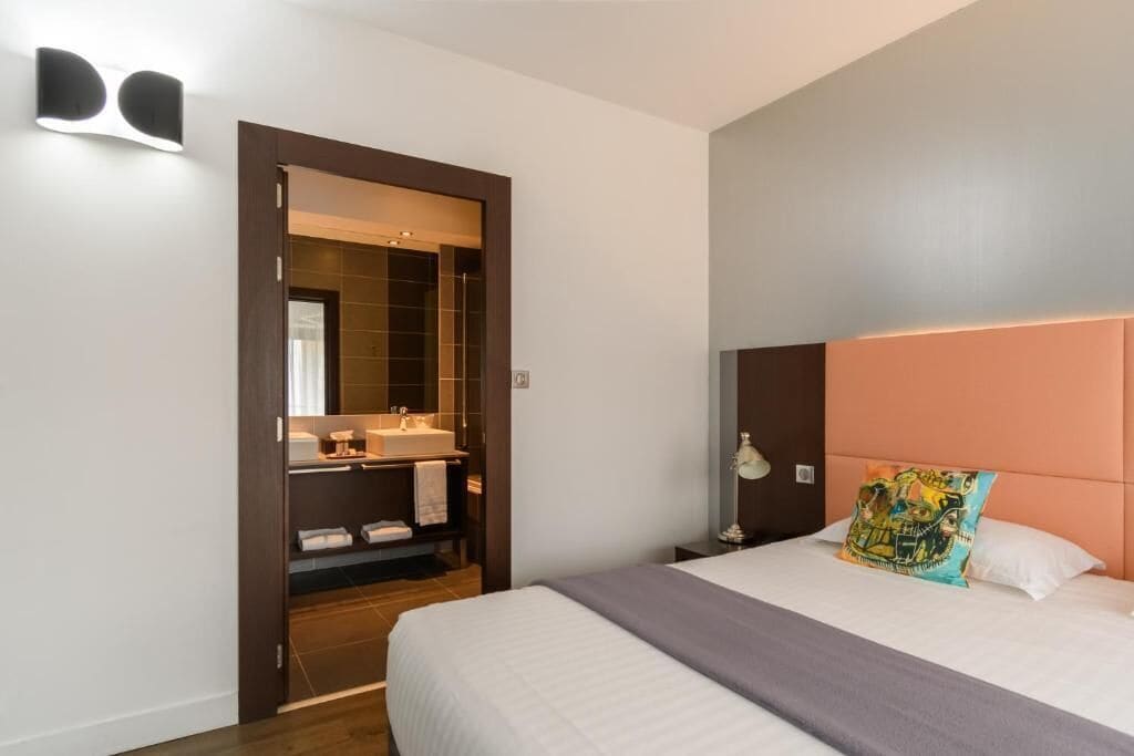 Une chambre d’hôtel comprenant un lit, une table de chevet, une commode, un miroir, une salle de bain avec un lavabo et un porte-serviettes, et une porte donnant accès à la salle de bain.