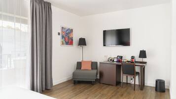 Una habitación de hotel moderna con piso de madera, un sofá gris, un escritorio con una silla, un televisor y un cuadro en la pared.