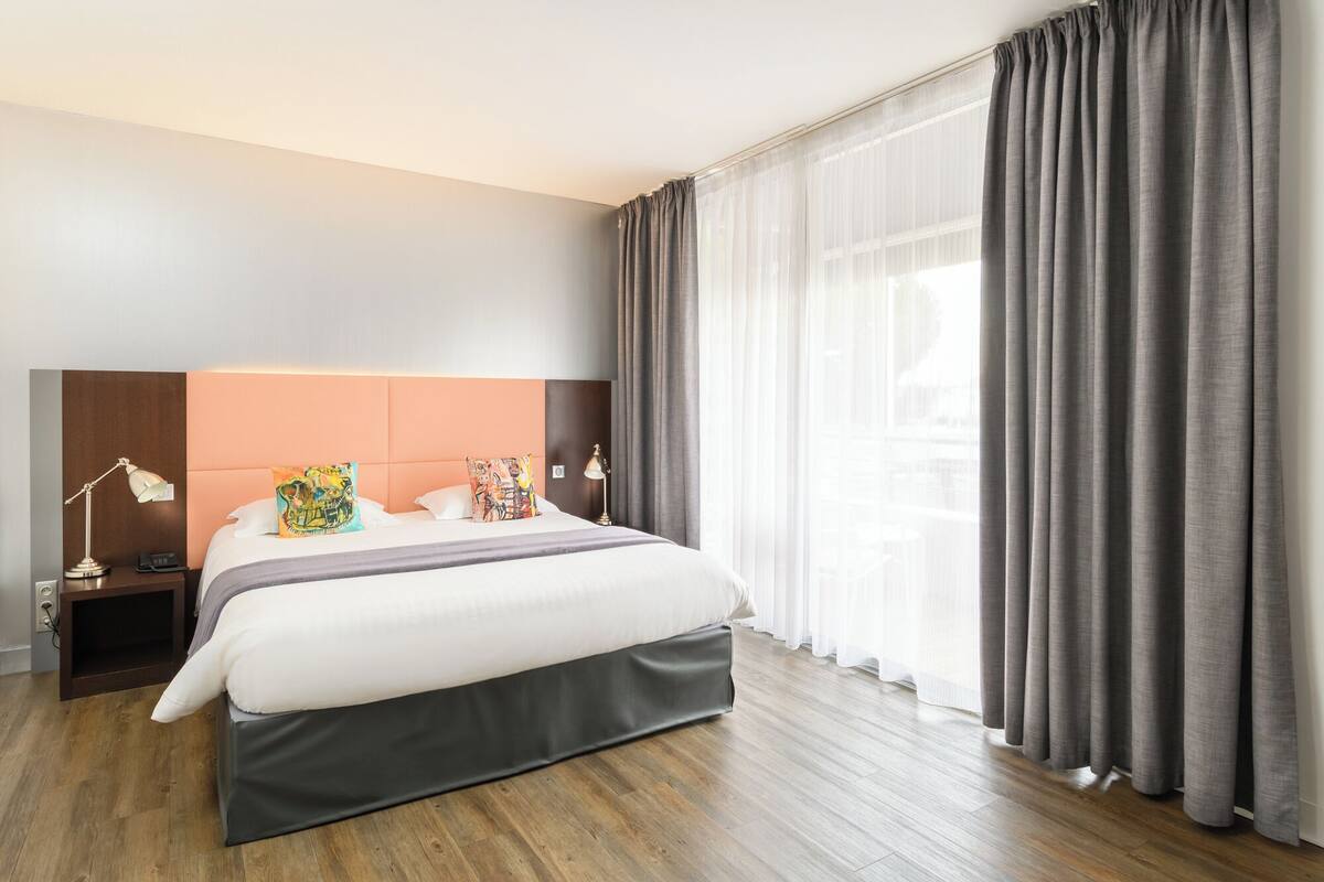 A modern hotel room with a large bed, a headboard, two bedside tables with lamps, and a view of the city through sheer curtains.