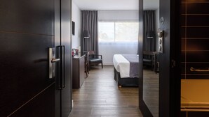 A modern hotel room with a bed, a desk, and a chair. There is a window with curtains and a door with a handle. - Hype Hotel (Biscarrosse)