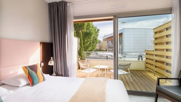 A modern hotel room with a large bed, a balcony with a view of a building and trees, and a small outdoor seating area. - Hype Hotel (Biscarrosse)