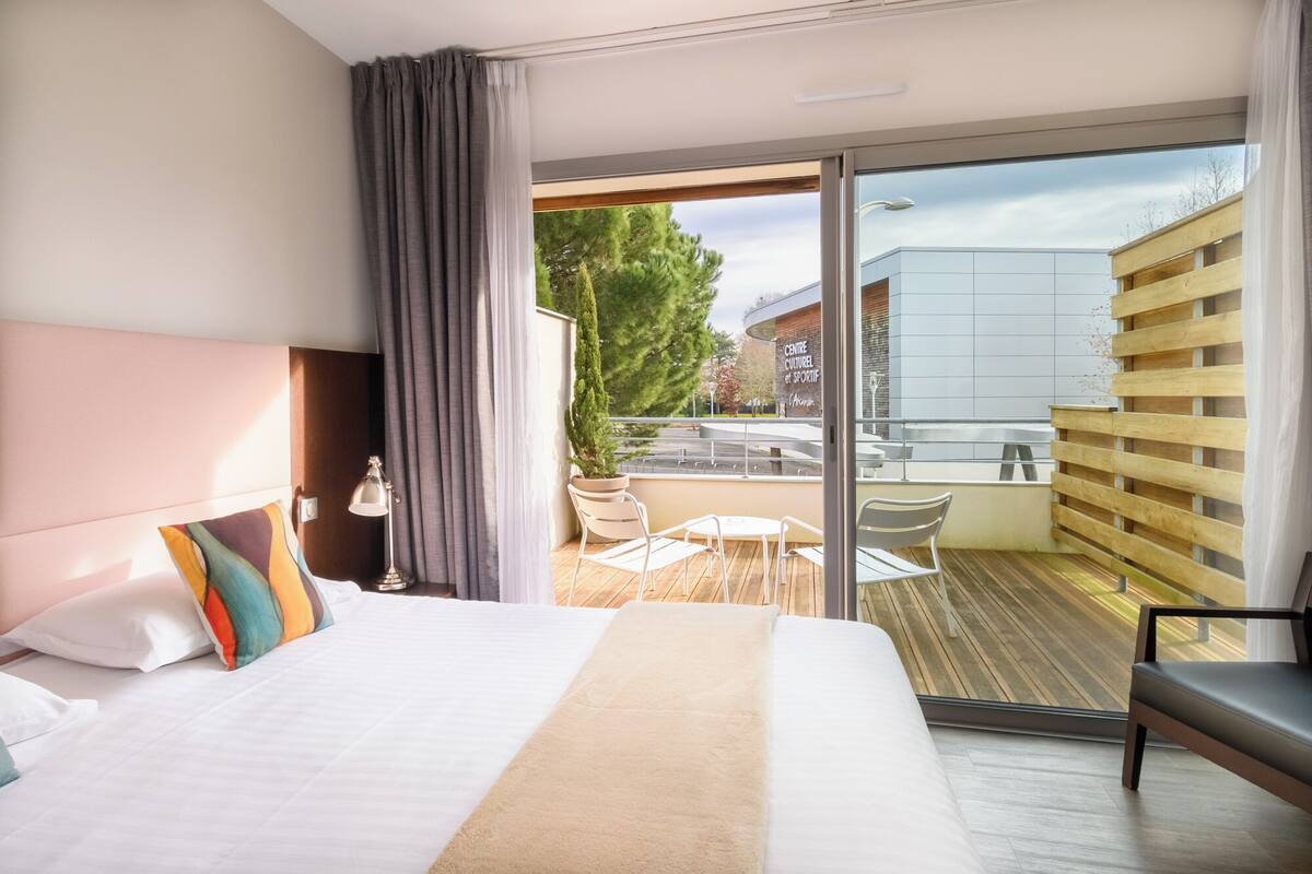 A modern hotel room with a large bed, a balcony with a view of a building and trees, and a small outdoor seating area.
