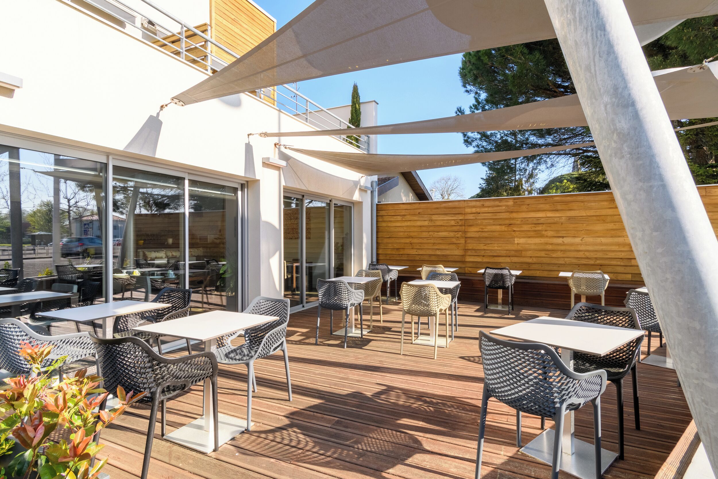 Een terras buiten met tafels en stoelen, een houten terras en een gebouw met grote ramen en een houten gevel.