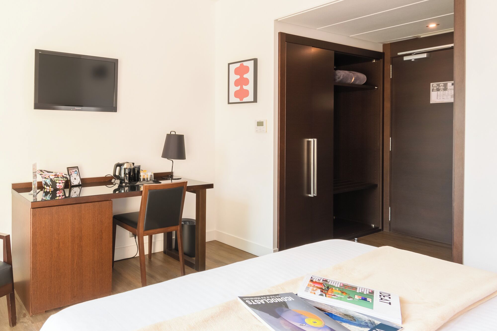 A hotel room with a bed, a desk with a chair, a lamp, a television, a wardrobe, and a magazine on the bed.